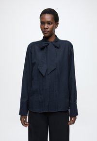 Tmavomodrá blúzka s dlhými rukávmi a viazaním na krku, vyrobená z textúrovanej tkaniny s vertikálnymi pruhmi a voľným strihom.