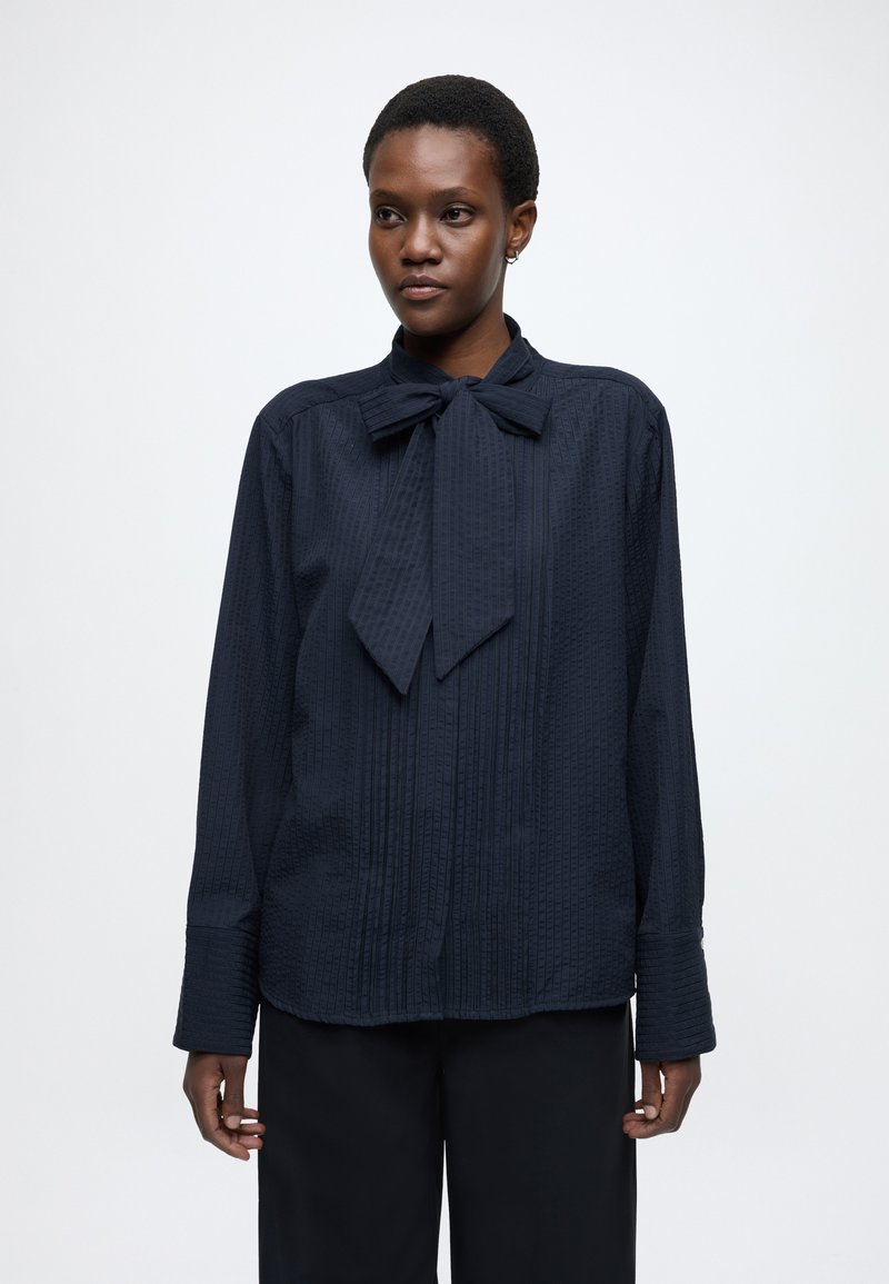 Tmavomodrá blúzka s dlhými rukávmi a viazaním na krku, vyrobená z textúrovanej tkaniny s vertikálnymi pruhmi a voľným strihom.