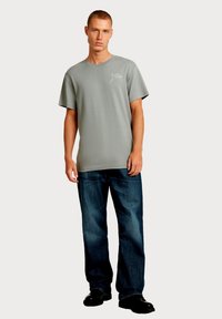 Grijs T-shirt met korte mouwen en een ronde hals, met een wit logo op de borst, gecombineerd met donkere rechte spijkerbroeken en zwarte schoenen.