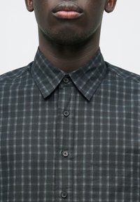 Tumehallist ruudulist musta nööbiga eest kinnitatav särk klassikalise kraega, läbipaistmatu kangast. Peen esitasku detail.