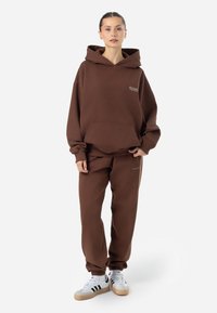 Felpa marrone con cappuccio e pantaloni della tuta abbinati, caratterizzati da una vestibilità comoda. La felpa presenta una tasca frontale e un logo. Il modello indossa sneakers bianche con strisce nere.