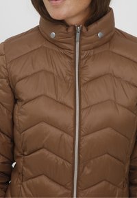 Femme portant une veste matelassée marron avec une fermeture éclair à l'avant et des boutons-pression sur le col montant.