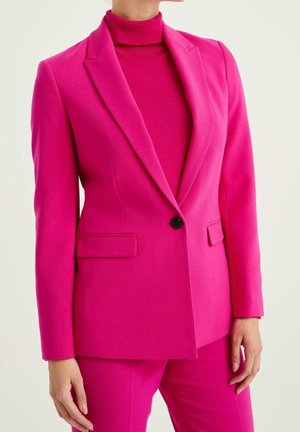 Kvinde iført en knaldrød skræddersyet blazer med en enkelt sort knap, matchende pink rullekrave og pink bukser mod en ensfarvet baggrund.