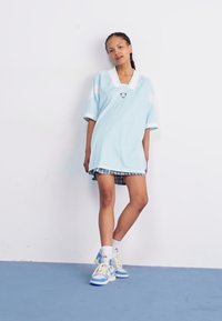 Lyseblå oversized polo kjole med hvide detaljer, prydet med et rundt logo. Båret over en ternet nederdel og kombineret med cremede og blå sneakers.
