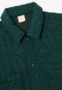 Σκούρο πράσινο κομμένο μπουφάν με κυκλικά σχέδια ραφής, μεταλλικά κουμπιά μπροστά, τσέπη στο στήθος και μπεζ ετικέτα XL μέσα στον γιακά.