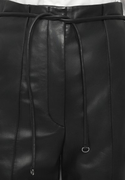 Κοντινό πλάνο από μαύρα δερμάτινα παντελόνια με ραμμένα ραφές και μια λεπτή δερμάτινη ζώνη που είναι δεμένη στη μέση πάνω από ένα λευκό πουκάμισο.