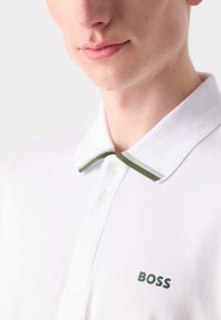 Balta polo krekls ar teksturētu apkakli un zaļiem akcentiem, kā arī zaļu "BOSS" logo. Klasisks griezums ar trim pogām ap kaklu.