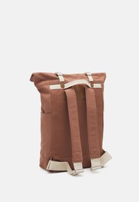 Bruine canvas rugzak met witte riemen, rol-top ontwerp, twee zijzakken en lichtgekleurde accenten. Duurzaam materiaal met gestikte details.