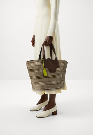 Personne en robe crème et chaussures blanches à petits talons tenant un grand sac fourre-tout tissé marron et beige avec des anses marron foncé et une étiquette vert citron.