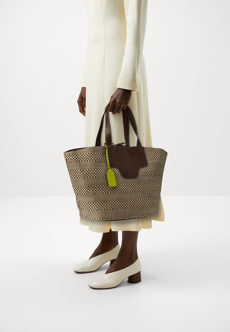 Personne en robe crème et chaussures blanches à petits talons tenant un grand sac fourre-tout tissé marron et beige avec des anses marron foncé et une étiquette vert citron.