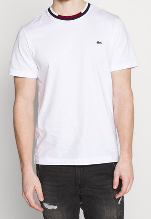 Mężczyzna w białym podkoszulku z krótkim rękawem i małym zielonym logo krokodyla na piersi oraz czarnych przetartych dżinsach.