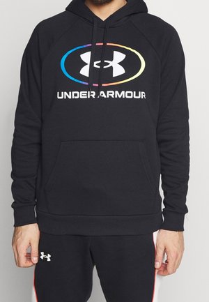 Homme portant un sweat à capuche noir Under Armour avec un logo ovale arc-en-ciel et un pantalon noir assorti avec un logo blanc et des bandes rouges sur les côtés.