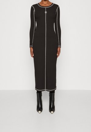 Kobieta ubrana w dopasowaną czarną sukienkę midi z długim rękawem i białymi kontrastowymi przeszyciami oraz czarne botki na kostkę ze spiczastym czubkiem i białymi końcówkami.