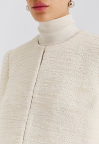 Krämfärgad jacka med struktur, rund halsringning och framknäppning. Bärs över en vit polotröja. Silvriga stiftörhängen är synliga.
