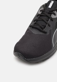 Zapato atlético negro con una parte superior de malla texturizada, diseño con cordones y suela de goma. Presenta un acento Reflectante y una puntera redondeada.