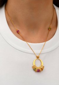 Collier en or avec un pendentif ovale orné de perles roses et vertes, chaîne texturée et accents décoratifs. Porté sur une chemise blanche.