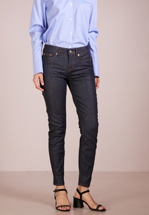 Persoon die donkere skinny jeans draagt, lichtblauw blouse met knopen en zwarte sandalen met blokhak en bandjes, staand tegen een effen achtergrond.