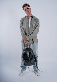Crna kožna ruksak s jednim gornjim ručkom, zatvaranjem na zip, i prednjim džepom. Model nosi sivi košulju s dugim rukavima i svijetle traperice.