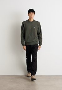 Felpa verde con collo tondo e logo, pantaloni neri e scarpe beige. Il modello è in piedi su uno sfondo bianco su un pavimento in cemento.