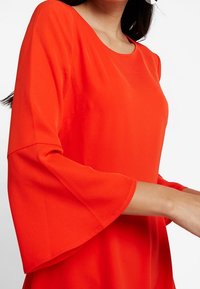 Blouse rouge à encolure ronde et manches cloche. Confectionnée dans un tissu lisse, arborant une couleur vive sans motifs ni ornements visibles.