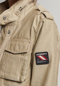 Khaki kurtka z przednimi kieszeniami, zamek błyskawiczny oraz naszywka na ramieniu z logiem "Superdry" w kolorach czerwonym, białym i niebieskim.