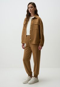 Bruine button-up jas met borstzakken, gecombineerd met bijpassende joggerpants met elastische manchetten, aangevuld met witte sneakers.