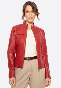 Rote Lederjacke mit Kragen, Reißverschluss vorne und zwei Brusttaschen. Kombiniert mit einem cremefarbenen Oberteil und beigen Hosen, der einen strukturierten Gürtel hat.