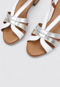 Sandales blanches avec des brides métalliques argentées, design ouvert au niveau des orteils, semelle intérieure marron et un petit talon empilé. Les brides sont texturées avec des accents de couture.