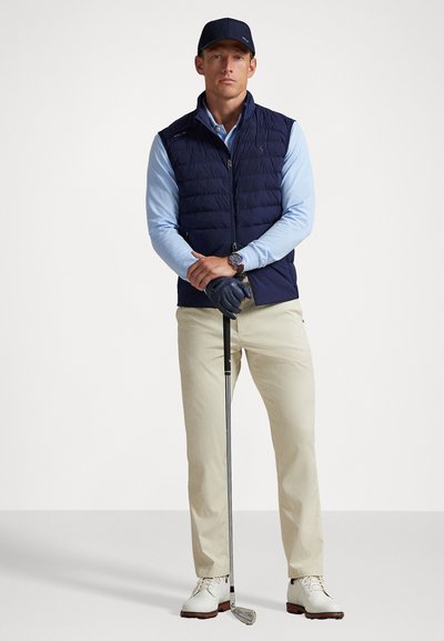 Man wearing navy cap, navy quilted vest, light blue long sleeve shirt, beige pants, white golf shoes, and glove, holding a golf club upright.