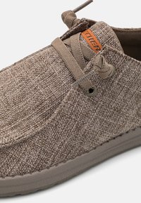 Brun texturerad sko med ovandel av vävd tyg, med elastiska snören, en ljusbrun logotypetikett och rund tåkonstruktion med flexibel sula.