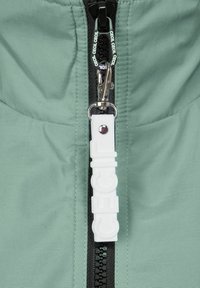 Chaqueta verde menta con cremallera frontal, tirador en blanco y negro, herrajes metálicos y detalles de textura acolchada.
