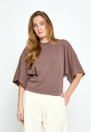 Frau mit langen hellbraunen Haaren, die ein lockeres, braunes kurzärmeliges Oberteil und hochtaillierte, off-white Hosen vor einem schlichten Hintergrund trägt.