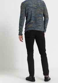 Blå och gul texturerad tröja med långa ärmar, kombinerad med svarta tajta byxor och svarta skor. Bakifrån syns tröjans sömdetaljer.