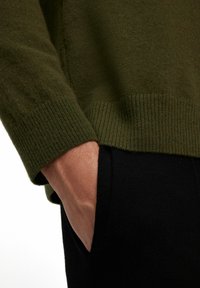 Maglione lavorato a maglia verde scuro con polsini a costine, mostrato con una mano appoggiata in una tasca di pantaloni neri con texture. Ingrandimento dei dettagli del tessuto.