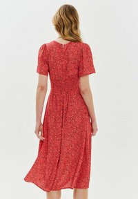 Červené květinové midi šaty s krátkými nabranými rukávy a smockovaným pasem. Vyznačují se jemnou, vzdušnou tkaninou a uvolněným siluetem. Detail knoflíků na zadní straně.