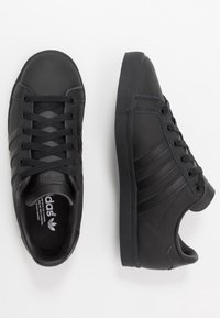 Mustat Adidas-lenkkarit naruilla, joissa on brändin kolme raitaa, yksi kenkä kuvattuna ylhäältä ja toinen sivuprofiilista valkoista taustaa vasten.