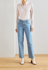 Camicia button-up a maniche lunghe con un motivo geometrico rosso e marrone ripetuto, abbinata a jeans dritti blu chiaro e stivaletti color crema.