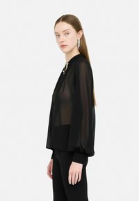 Zwarte, doorschijnende blouse met lange mouwen, een hoge hals en geplooide manchetten, met decoratieve hardware bij de kraag.