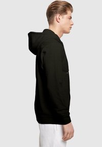 Sort hættehætte med en forhåndslomme, ribbede manchetter og en afslappet pasform. Stoffet har en glat tekstur. Modellen vist bærer hvide bukser.