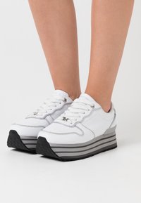 Bílé platformové tenisky se šedým pruhováním, koženým svrškem, šněrováním a stříbrnými akcenty na jazyku a po stranách. Černá gumová podešev.