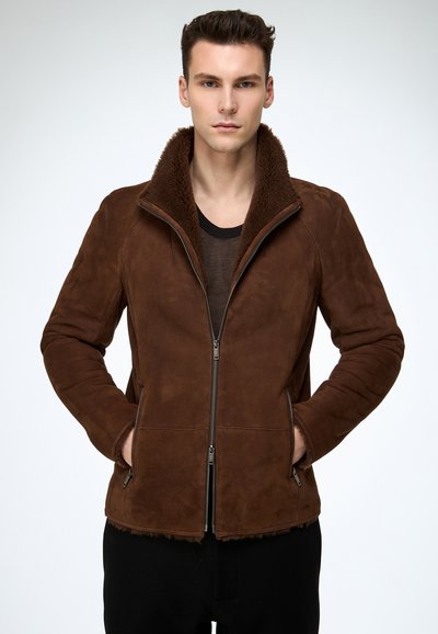 Mann trägt eine braune Reißverschluss-Lammfelljacke mit hochgestelltem Kragen, Hände in den Taschen, steht vor einem einfarbigen hellen Hintergrund.