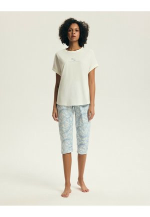 Kobieta stojąca boso, ubrana w krótki, kremowy top z krótkim rękawem oraz jasnoniebieskie spodnie 7/8 z białym wzorem liści na jednolitym tle.