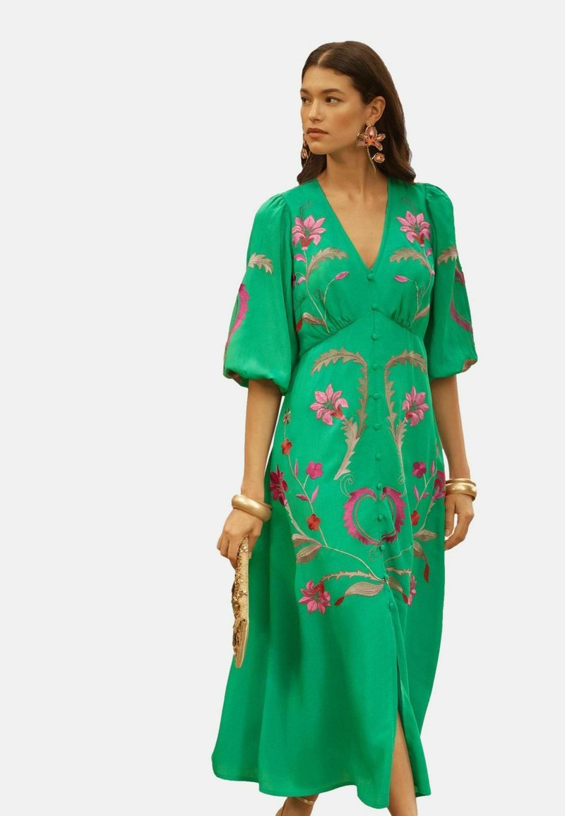 Woman wearing a green dress with pink floral embroidery, puffed sleeves, holding a gold clutch, and accessorized with large earrings and bangles.
