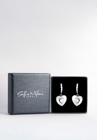 Silverfärgade hjärtformade örhängen med klara ädelstenar, placerade i en svart presentask med präglad logotyp i vitt.
