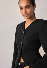 Blazer nero con scollatura a V profonda, tre bottoni metallici e maniche strutturate. Il tessuto ha una texture liscia e una vestibilità su misura.