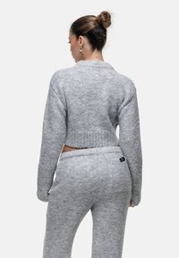 Vrouw met een knot in haar haar draagt een grijze fuzzy cropped trui en bijpassende broek, met haar rug naar de camera op een plain witte achtergrond.
