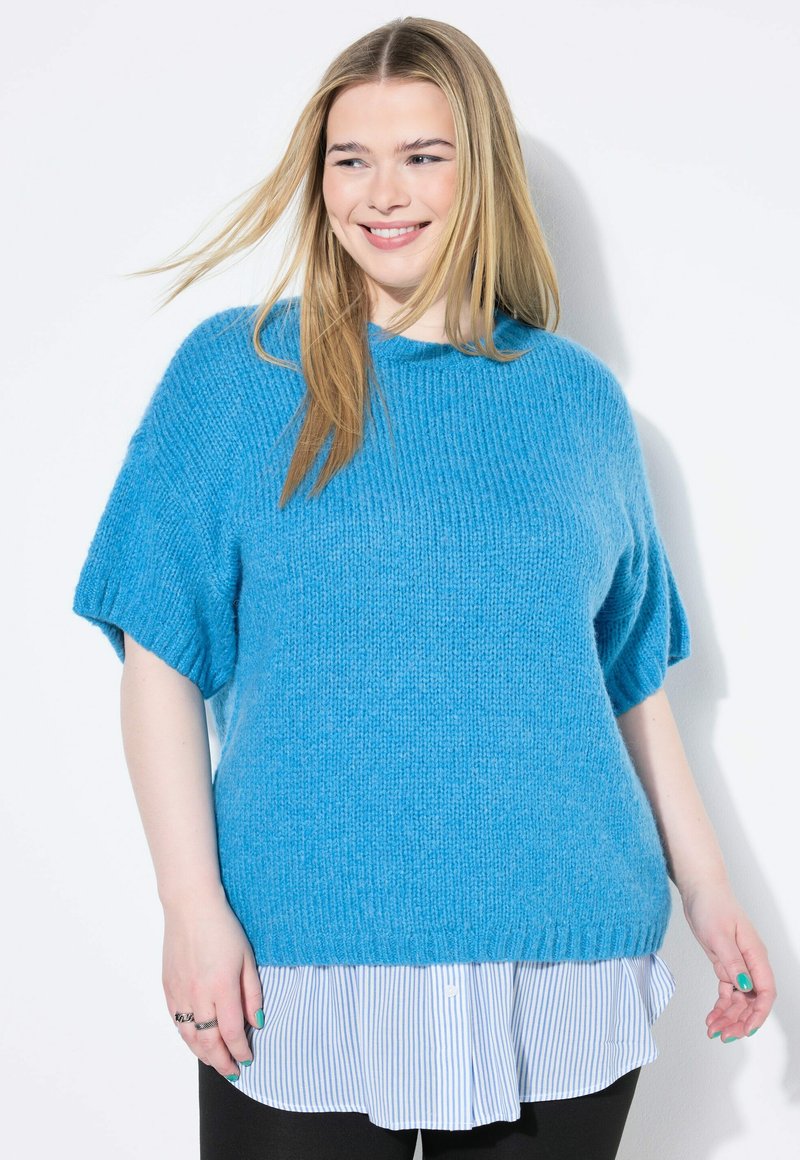 Ljusblå stickad tröja med korta ärmar, ribbad halsringning, lagd över en vit och blårandig skjorta. Slät textur och avslappnad passform.