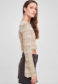 Beige, strikket crop topp med horisontale striper og åpningsmønstre. Langermede ermer med utbrettede mansjetter. Svart shorts brukt under.