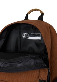 Innenseite eines braunen Cordura-Rucksacks mit schwarzem Futter, einem beschrifteten Etikett mit der Aufschrift "DIESE TASCHE GEHÖRT ZU:" und einem Reißverschlussfach.