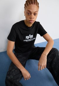 T-shirt en coton noir avec le logo blanc "Alpha Industries", associé à un pantalon noir arborant un motif floral discret, assis contre un fond clair.